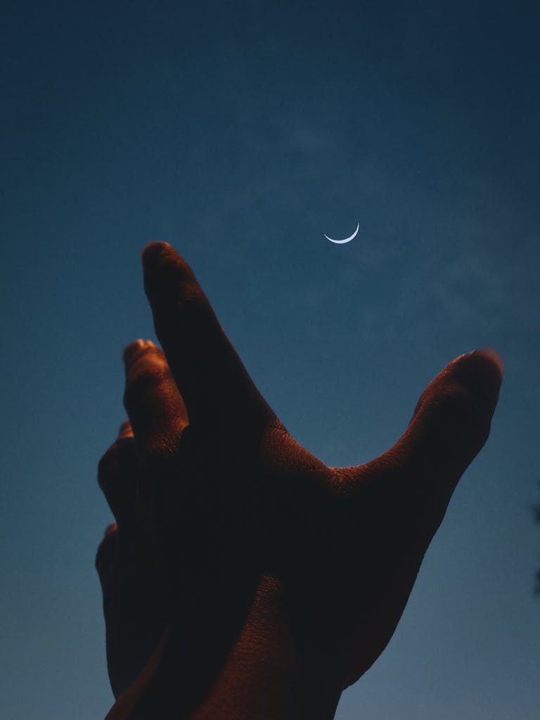 Silhouette of a hand reaching towards a crescent moon in a dark night sky.